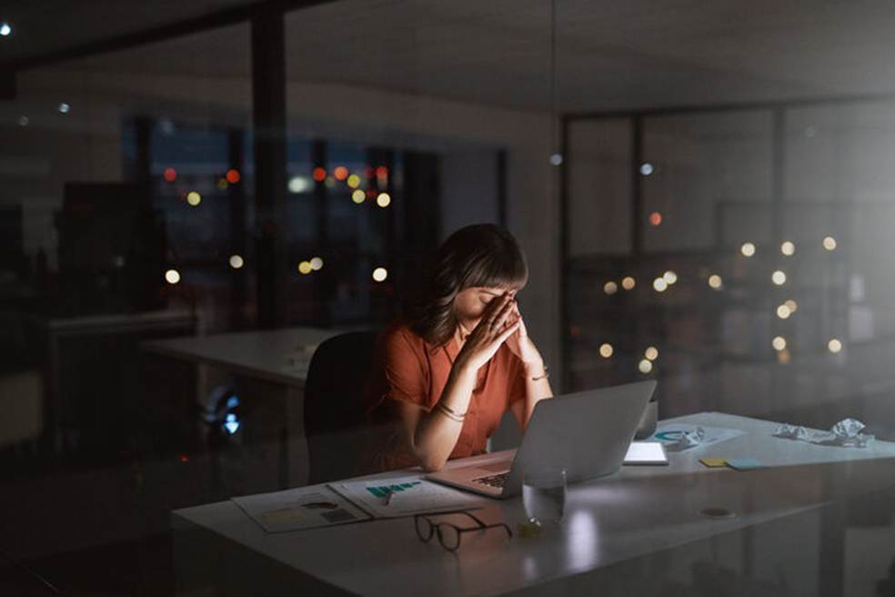 A woman at a desk, working late, with her head in her hands. Papers, glasses, and a glass of water sit on the desk.